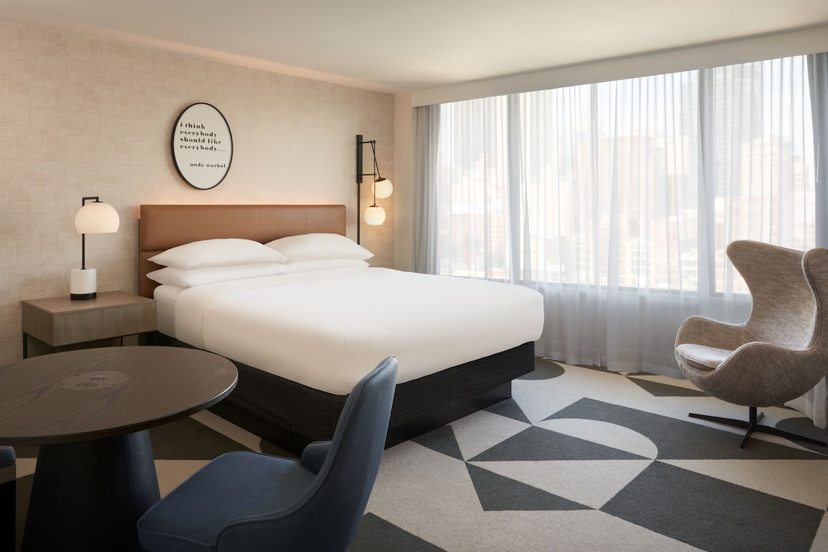 Habitación de hotel moderna con una cama grande, un escritorio, una silla y vistas de la ciudad a través de cortinas translúcidas.