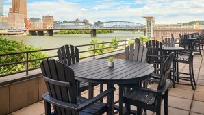 Una terraza con mesas redondas y sillas Adirondack, con vista a un río que tiene un puente y edificios de la ciudad al fondo.
