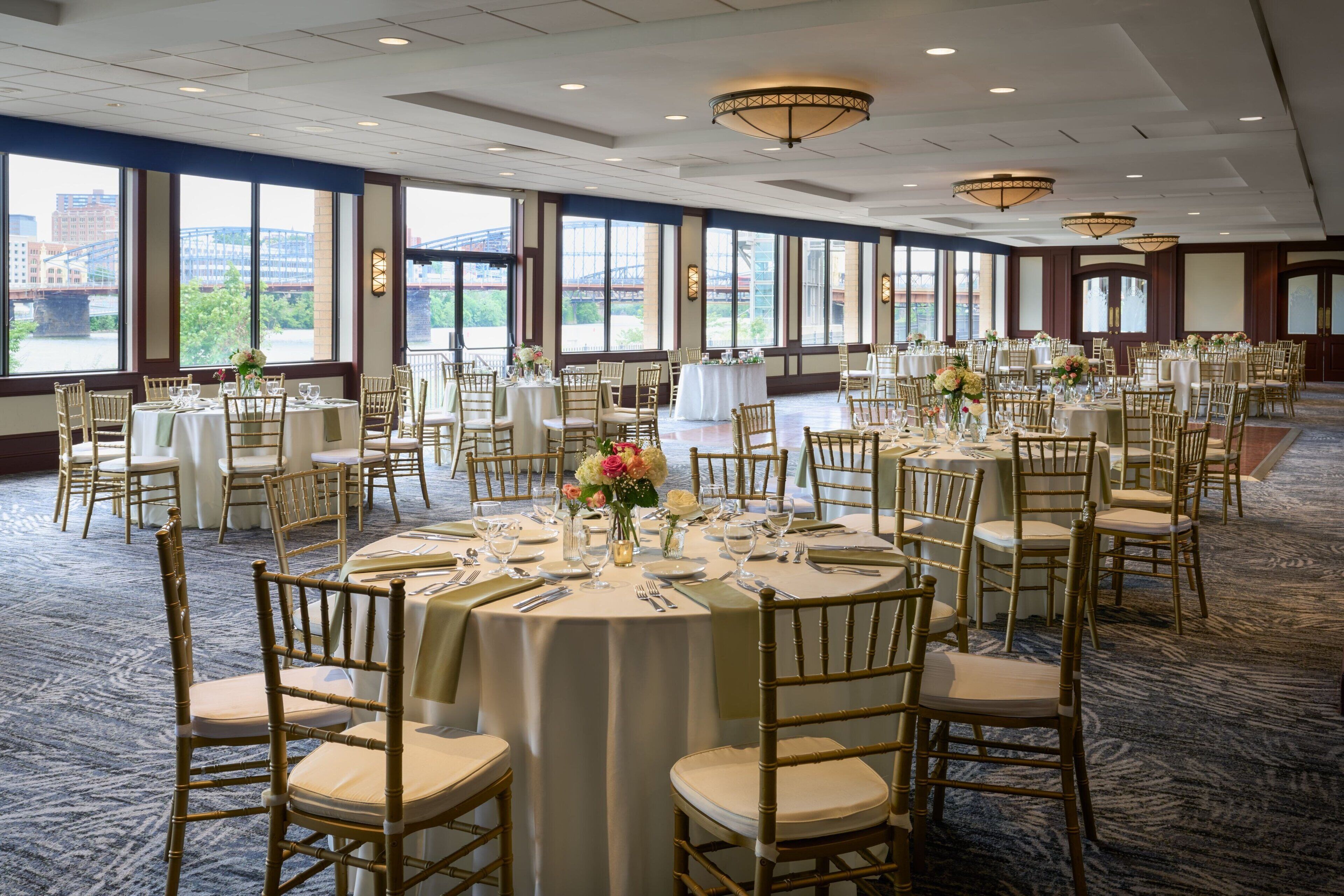 Une salle de banquet avec des tables rondes, des chaises dorées et de grandes fenêtres donnant sur un pont et des immeubles.