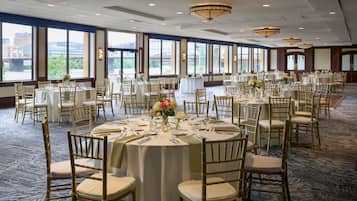 Une salle de banquet avec des tables rondes, des chaises dorées et de grandes fenêtres donnant sur un pont et des immeubles.