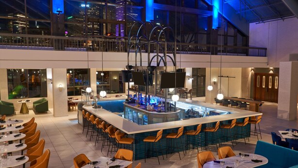 Zona de bar moderna con una isla central, asientos alrededor y vistas de la ciudad a través de amplios ventanales.
