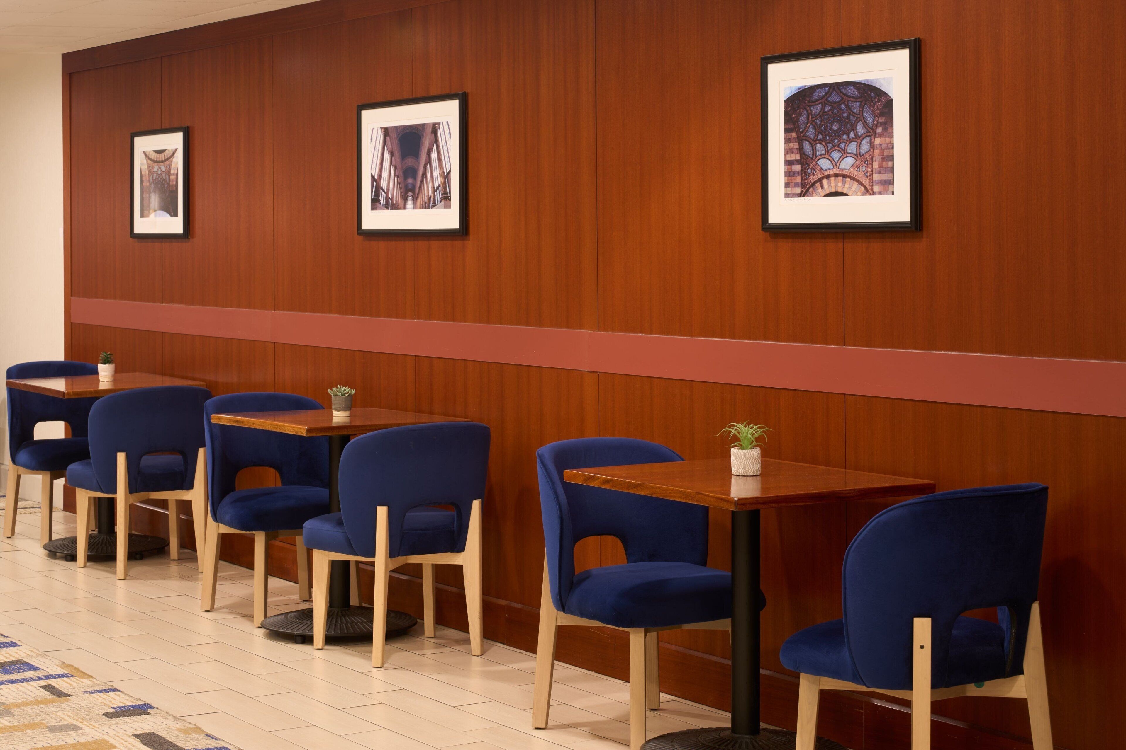 A hotel lobby with wooden panel walls, framed pictures, blue upholstered chairs, and wooden tables.