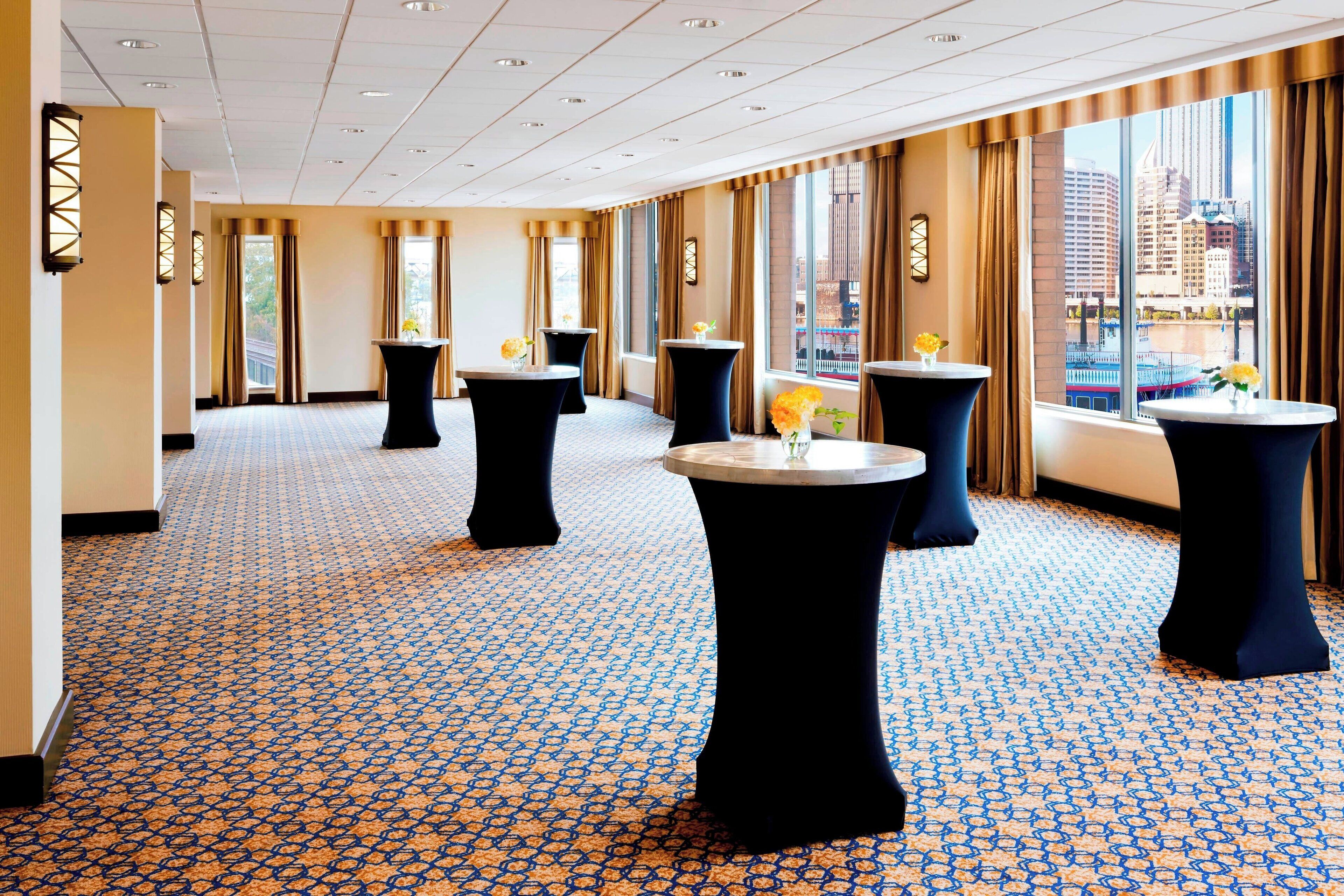 A hotel lobby with patterned carpet, round tables with vases, and large windows with curtains.