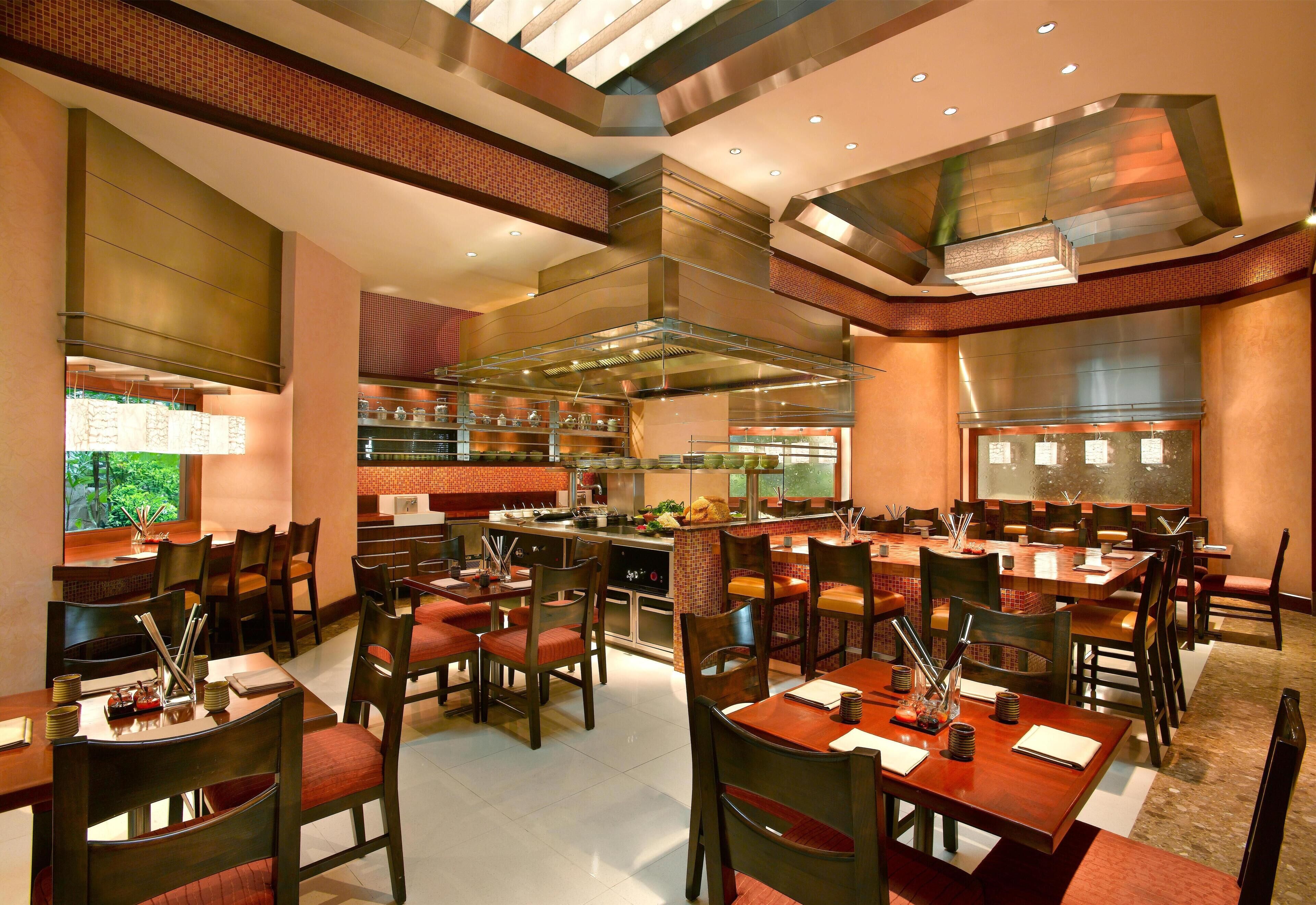 Um restaurante amplo com várias mesas e cadeiras, uma área de cozinha central e vista para o exterior através de grandes janelas.