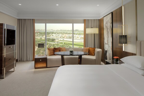 A hotel room with a large bed, a sofa, a small round table, a TV, and a view of a cityscape.