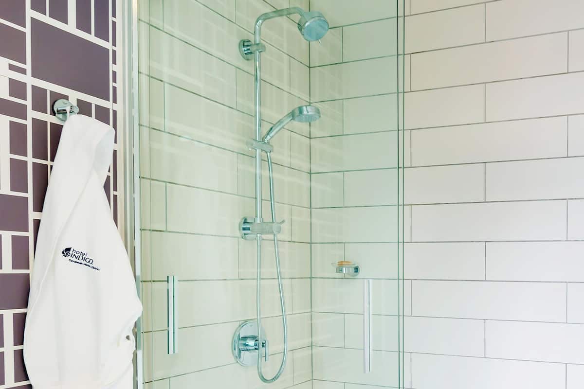 Eine in Glaswände gefasste Dusche mit Chromarmaturen, ein weißes Handtuch an der Wand und eine Wand mit Kachelfliesen in Rasteroptik.