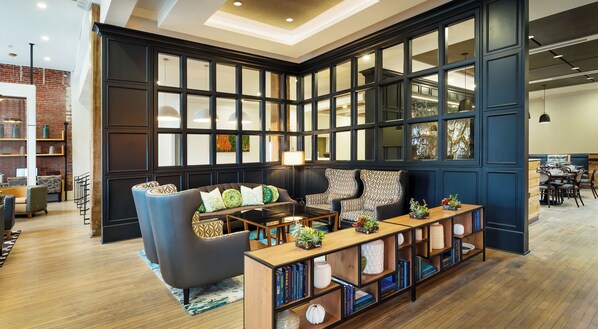 Relax in a cozy lounge area with a comfortable sofa set, coffee table, and bookshelf, surrounded by large windows and an exposed brick wall.