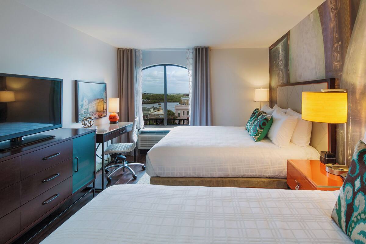 A hotel room with two beds, a desk, a TV, and a view of the outdoors.