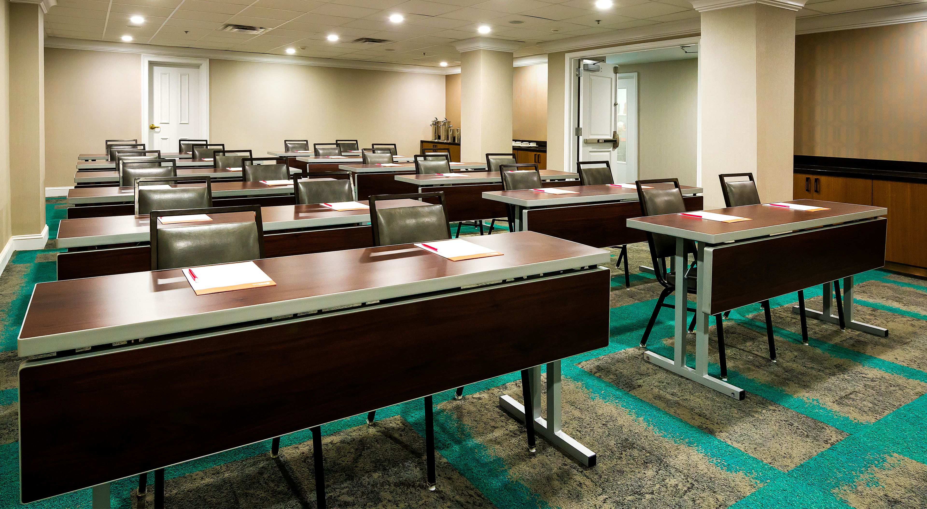A conference room with multiple tables and chairs, carpeted floor, and a door in the background.