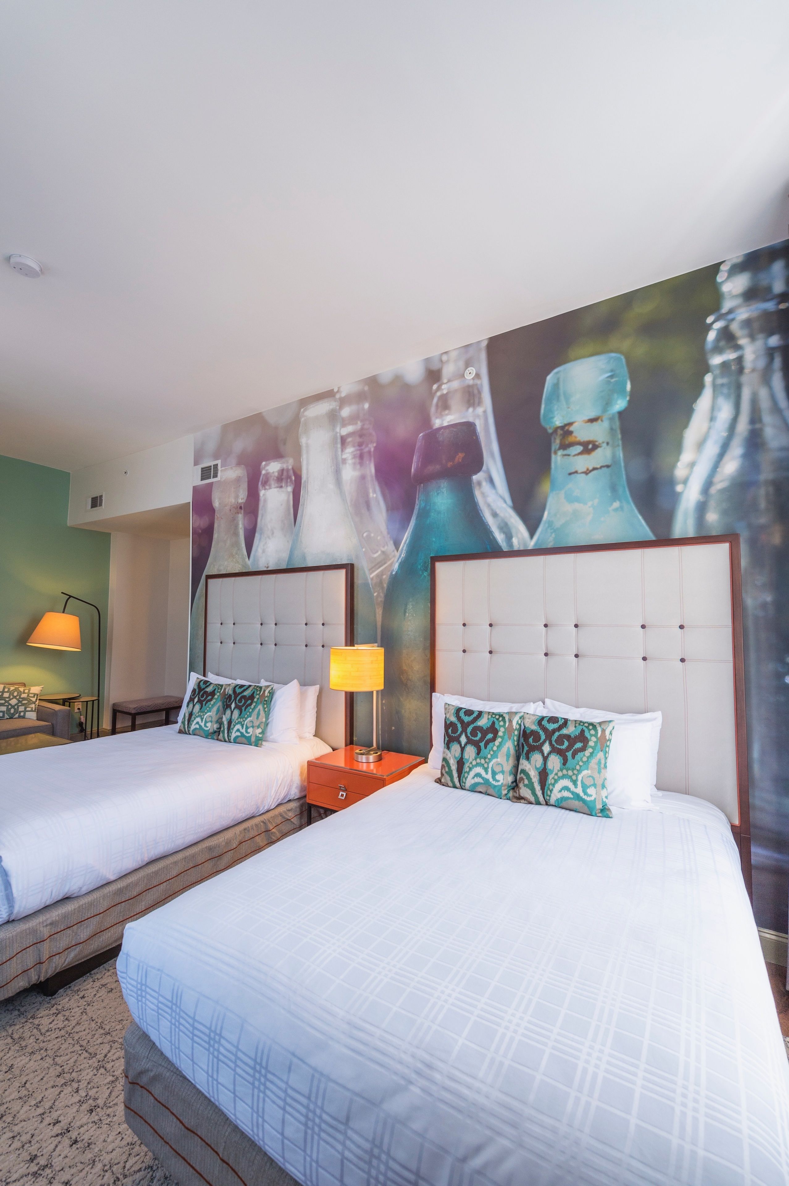 A hotel room with two beds, a large wall mural of bottles, a bedside table with a lamp, and a small desk with a chair.