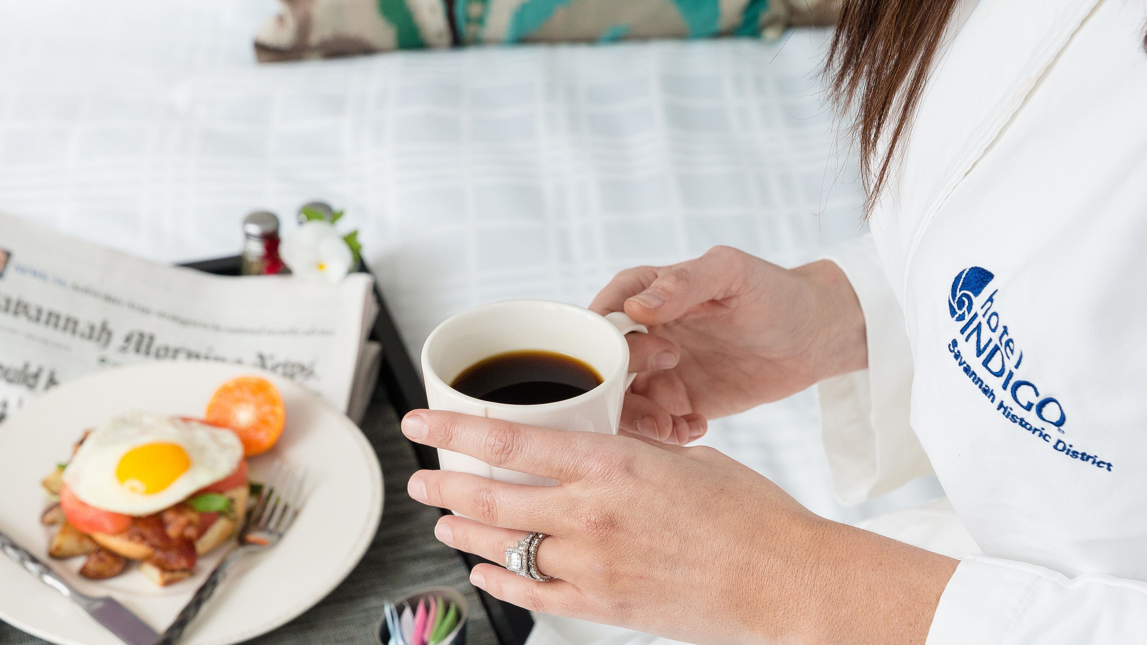 A person holding a coffee mug, a newspaper, and a plate with breakfast.