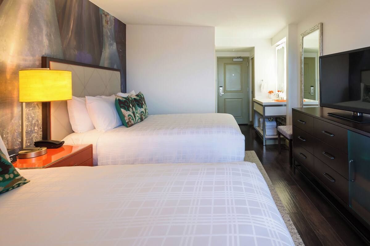 A hotel room with two beds, a TV, a desk with a lamp, and a bathroom visible in the background.