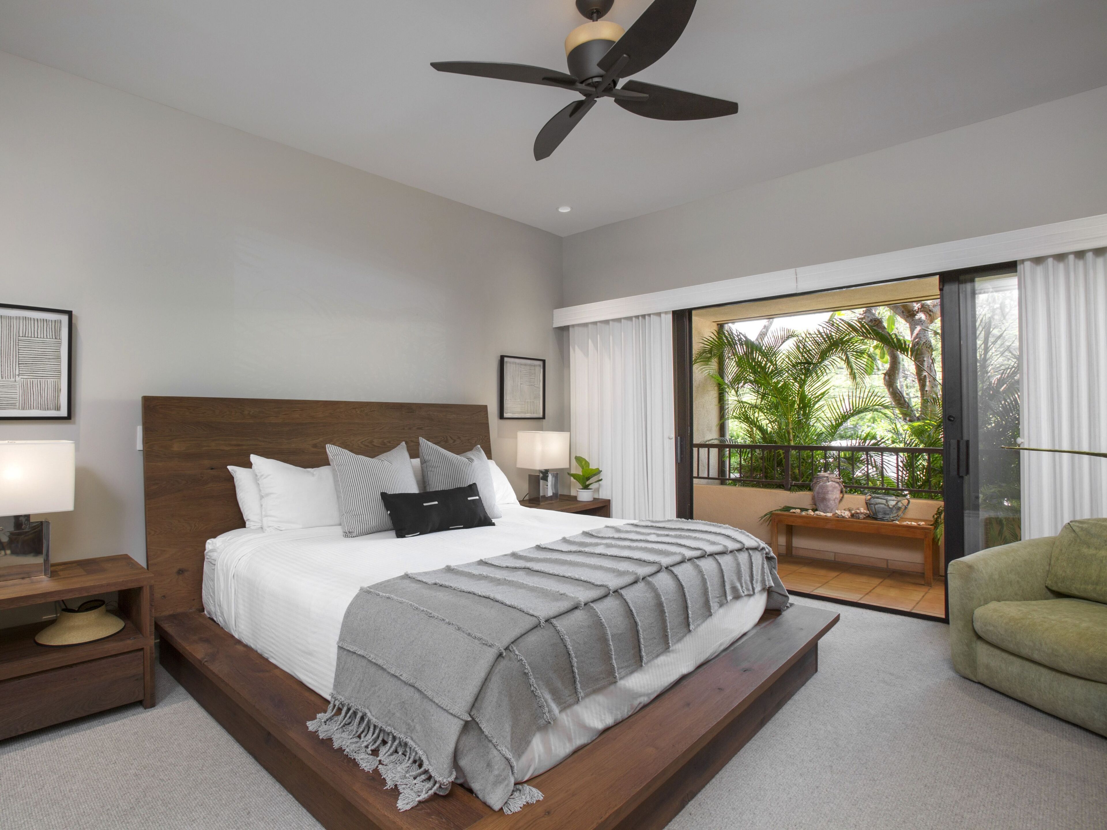 A modern bedroom with a large bed, a ceiling fan, a nightstand with a lamp, a green armchair, and a balcony with plants.
