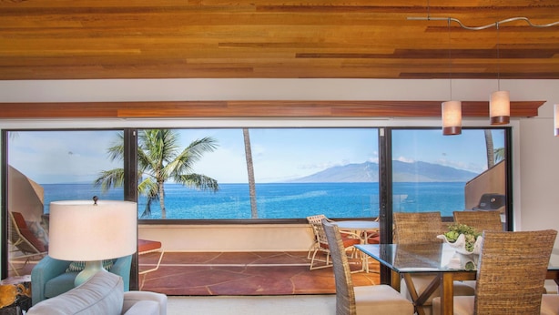 Una habitación con un ventanal grande con vistas al océano, muebles de mimbre, un sofá y una mesa con una lámpara.