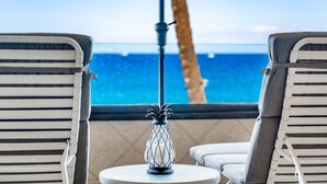 Un espacio al aire libre iluminado por el sol con vista al océano, con dos sillones de descanso y una mesa con un candelabro decorativo.