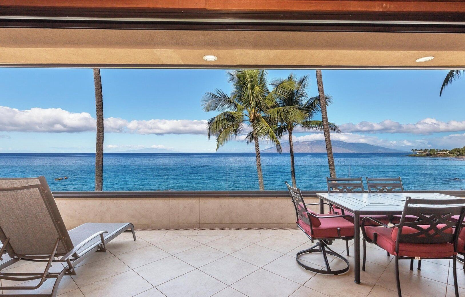 A room with a large window overlooking the ocean, a dining table with chairs, and a lounge chair.