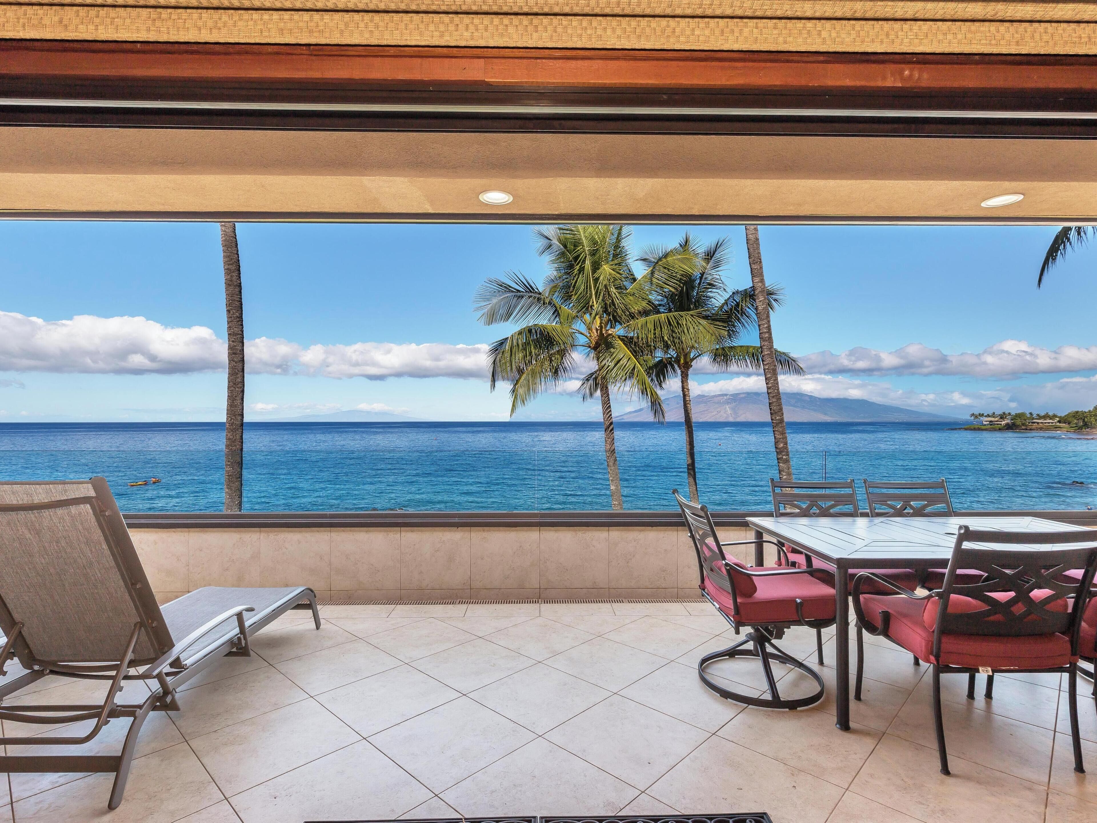 A balcony with a view of the ocean, palm trees, and a table with chairs.