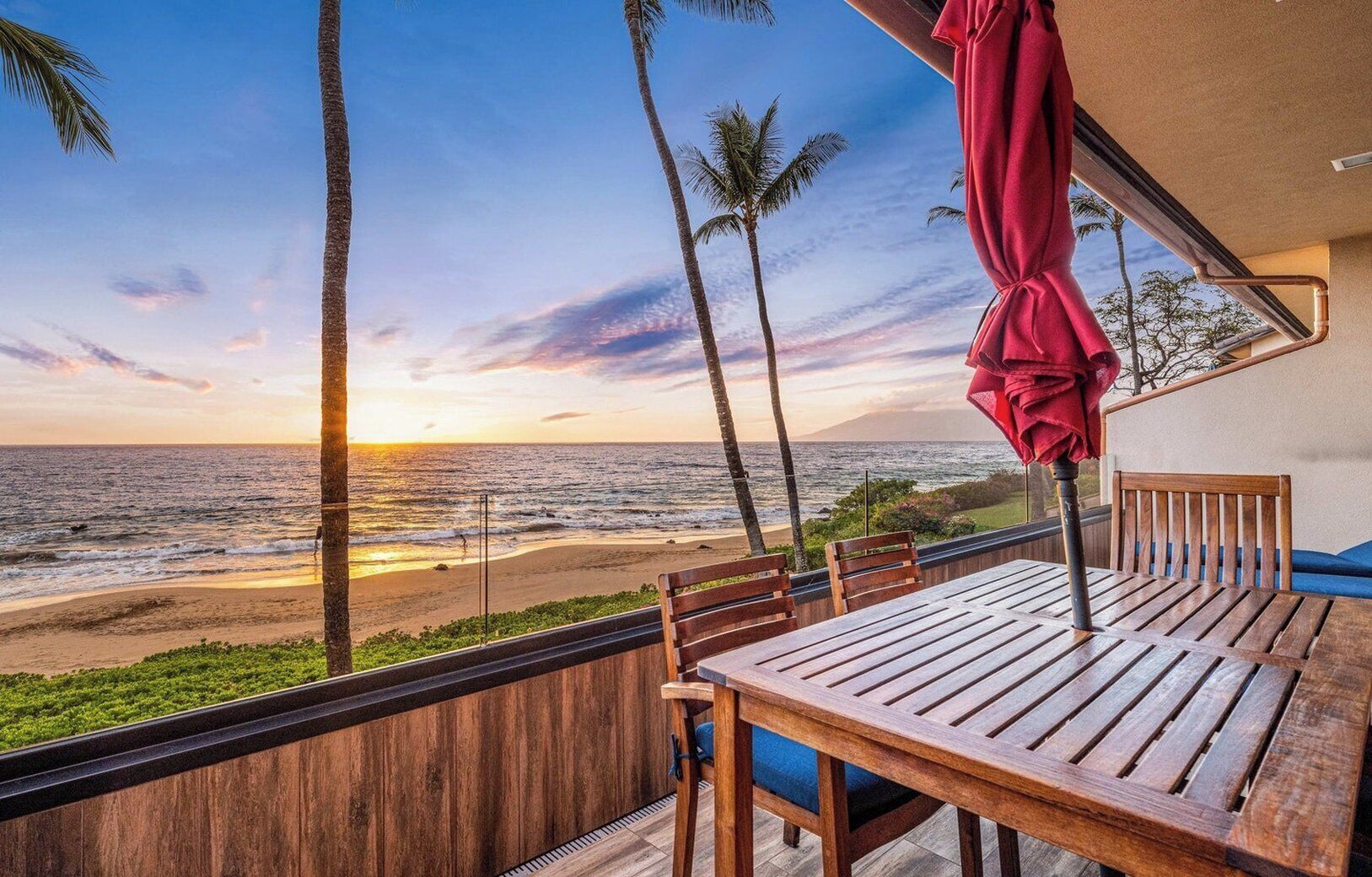 A wooden deck with a table and chairs, overlooking a beach with palm trees and the ocean at sunset.