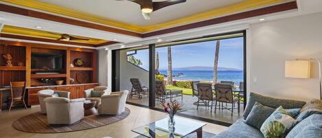 A spacious living room with a large sofa, a TV, and a dining area with chairs. There is a view of the ocean through large sliding doors.