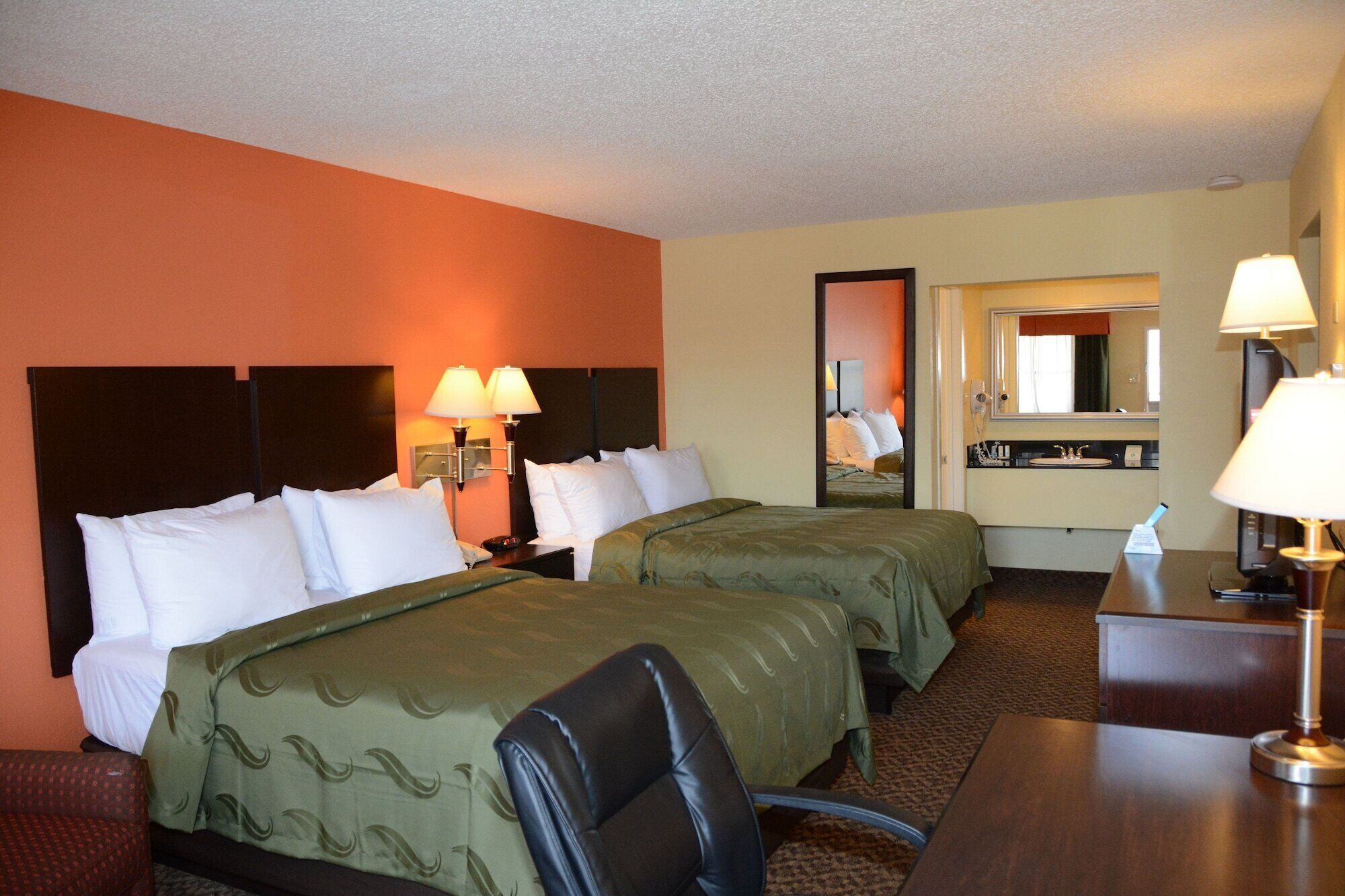 A hotel room with two beds, a desk, a chair, a mirror, and a bathroom visible through an open door.