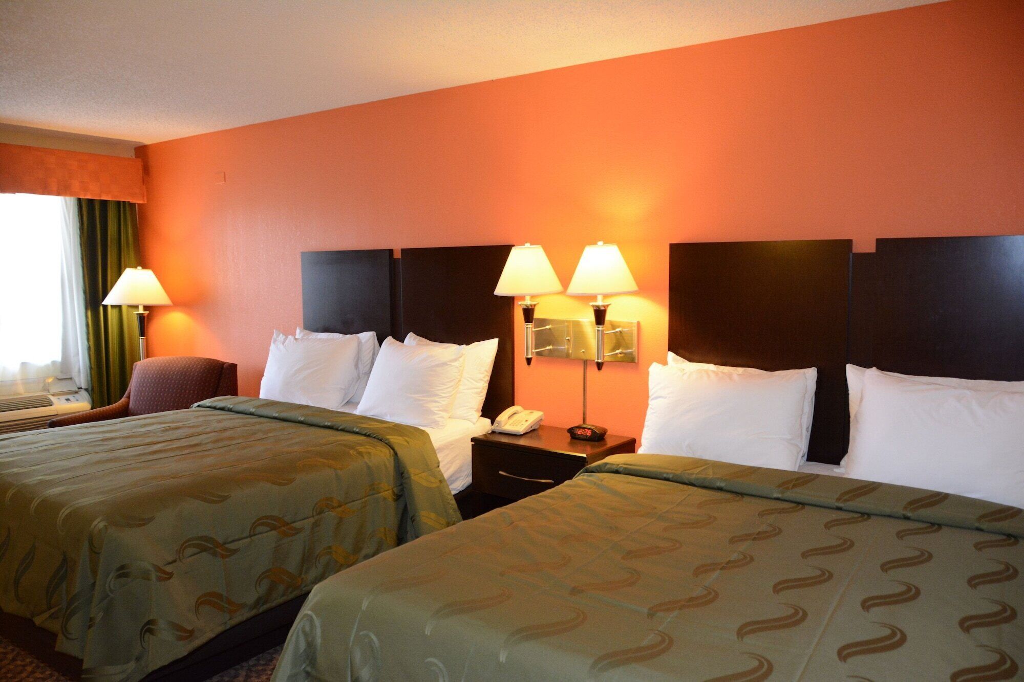 A hotel room with two beds, a nightstand with a phone and alarm clock, and two wall-mounted lamps.