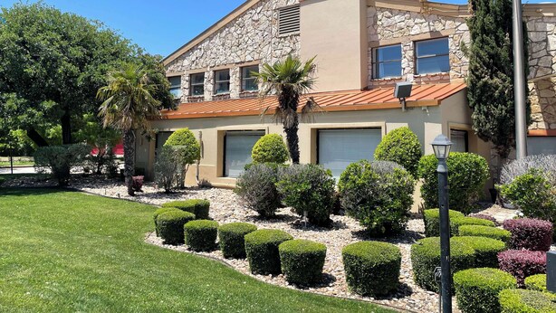 Une maison à deux étages avec une façade en pierre, un toit recouvert de tuiles rouges et un jardin bien entretenu agrémenté de haies et d’un palmier.