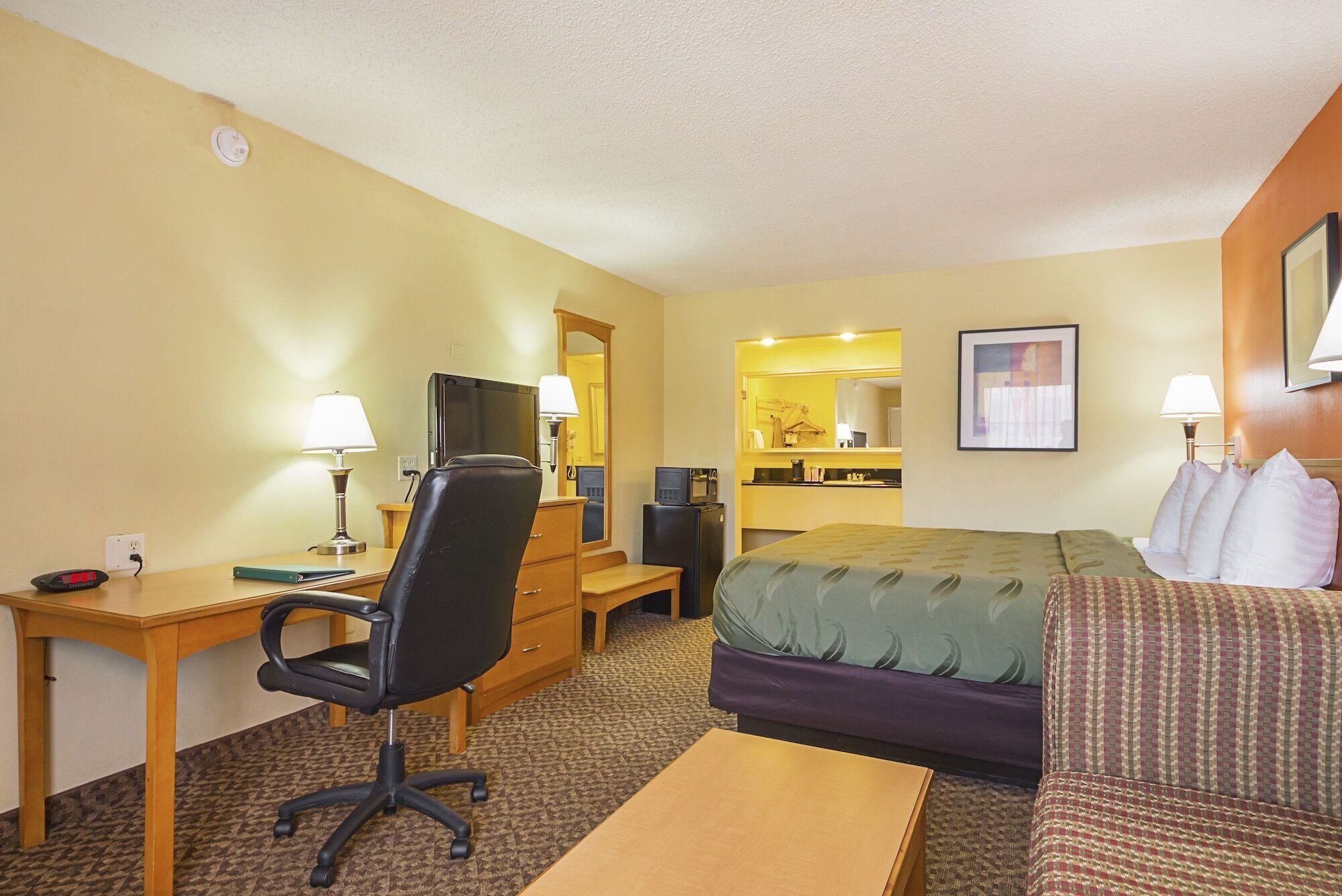 Una habitación de hotel con cama, escritorio con computadora, silla, televisión y kitchenette pequeña.