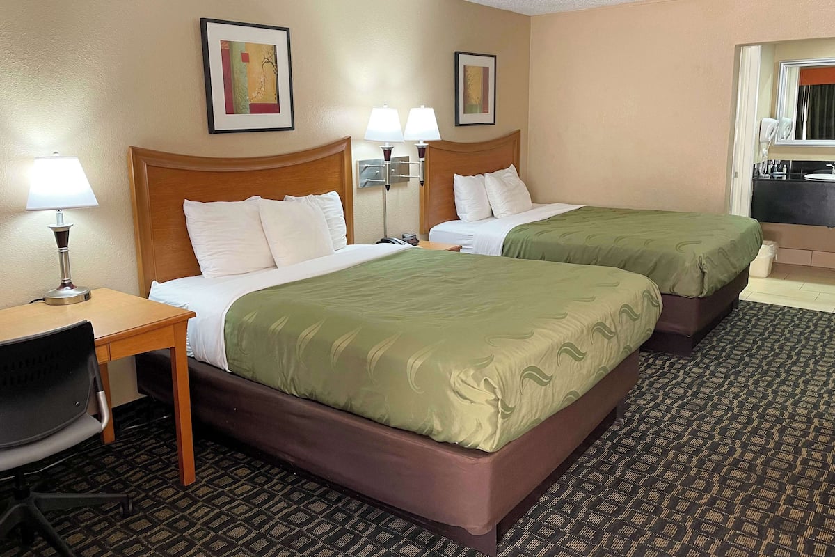 A hotel room with two beds, a desk, a chair, and a bathroom visible through an open door.