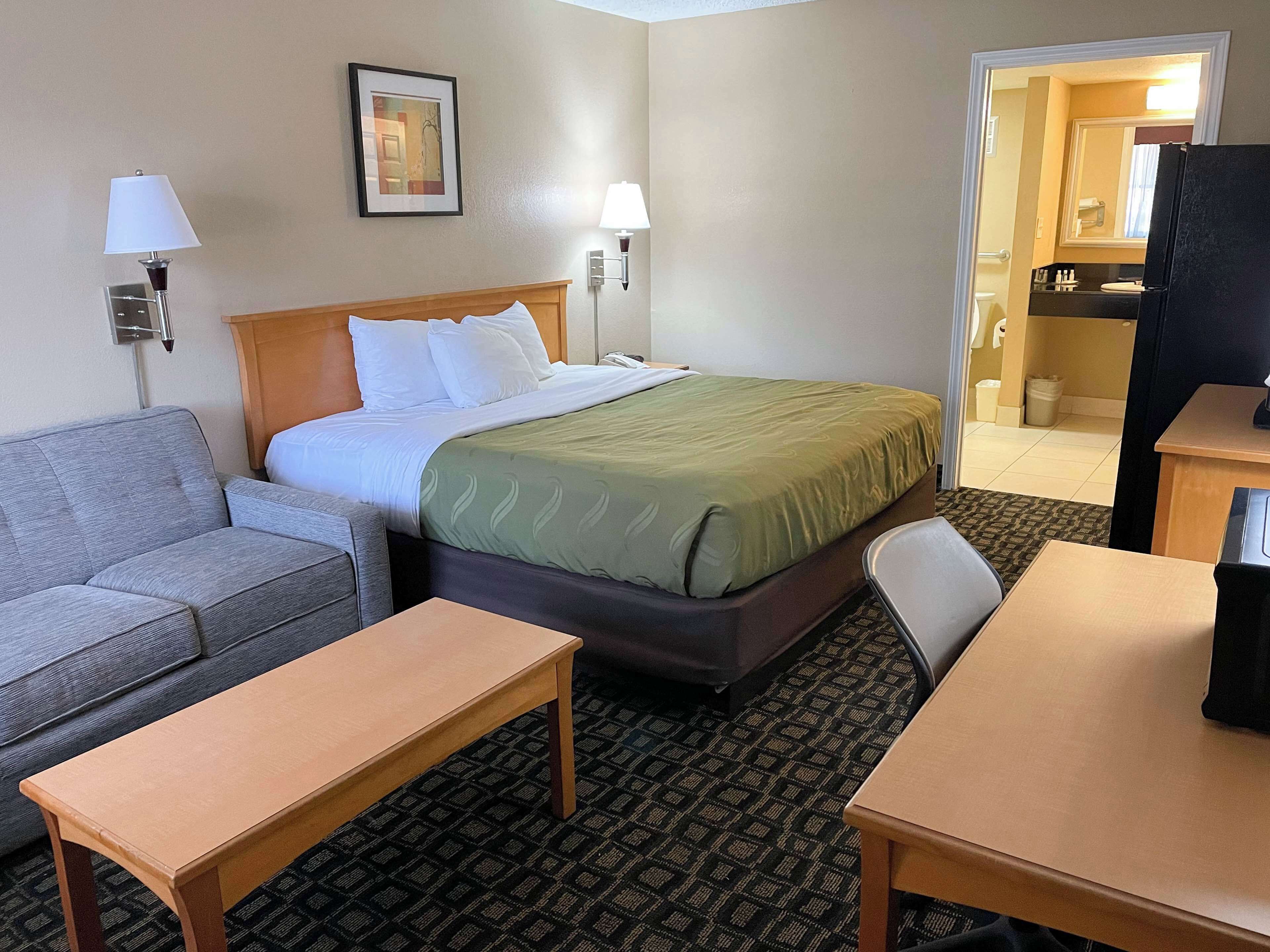 Habitación de hotel con cama, sofá, mesa y silla. Se ve un baño por una puerta abierta.