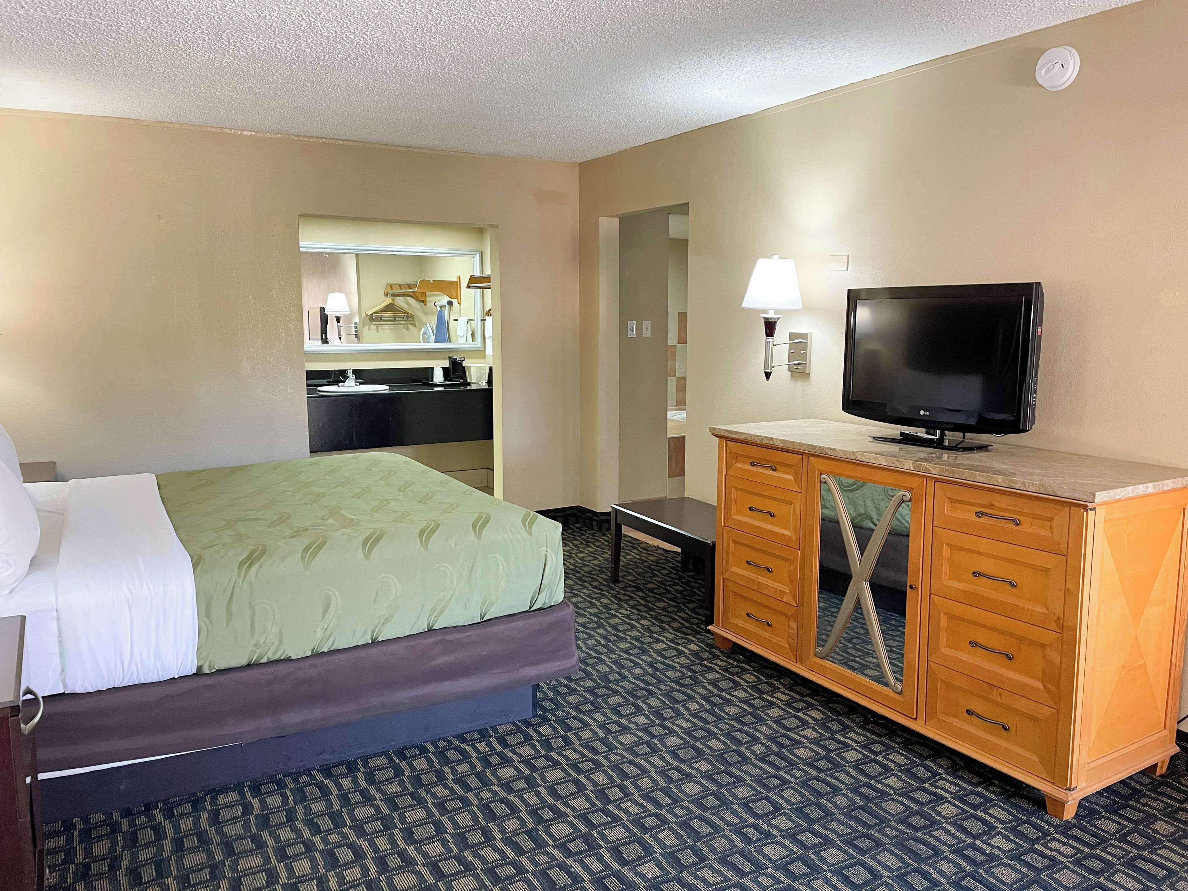 A hotel room with a bed, a TV on a wooden cabinet, a desk, and a bathroom visible through an open door.