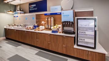 A hotel breakfast area with a self-serve station, including yogurt, eggs, and milk dispensers, and a sign indicating the start of breakfast.