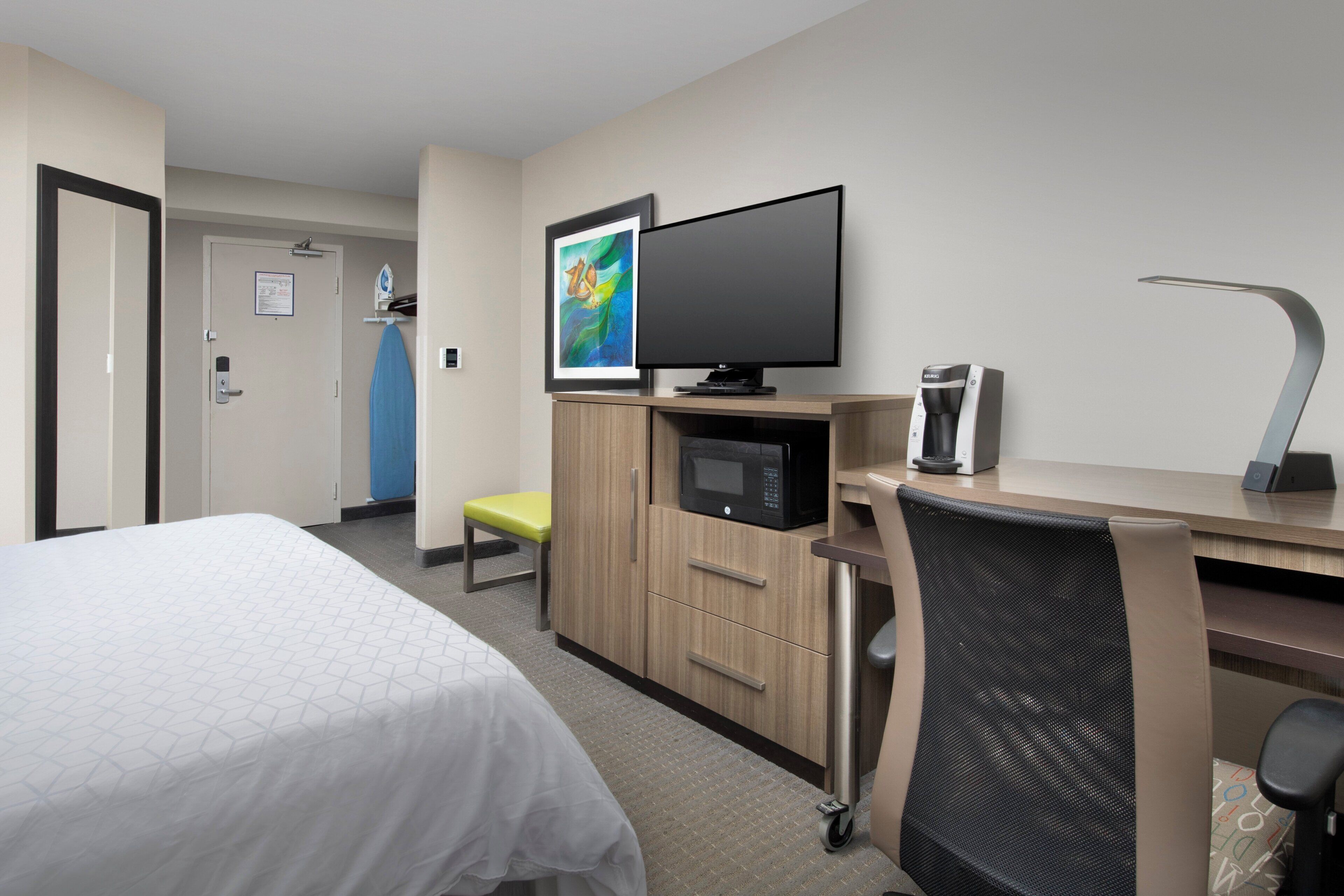A hotel room with a bed, a desk with a computer, a flat-screen TV, a microwave, and a coffee machine.