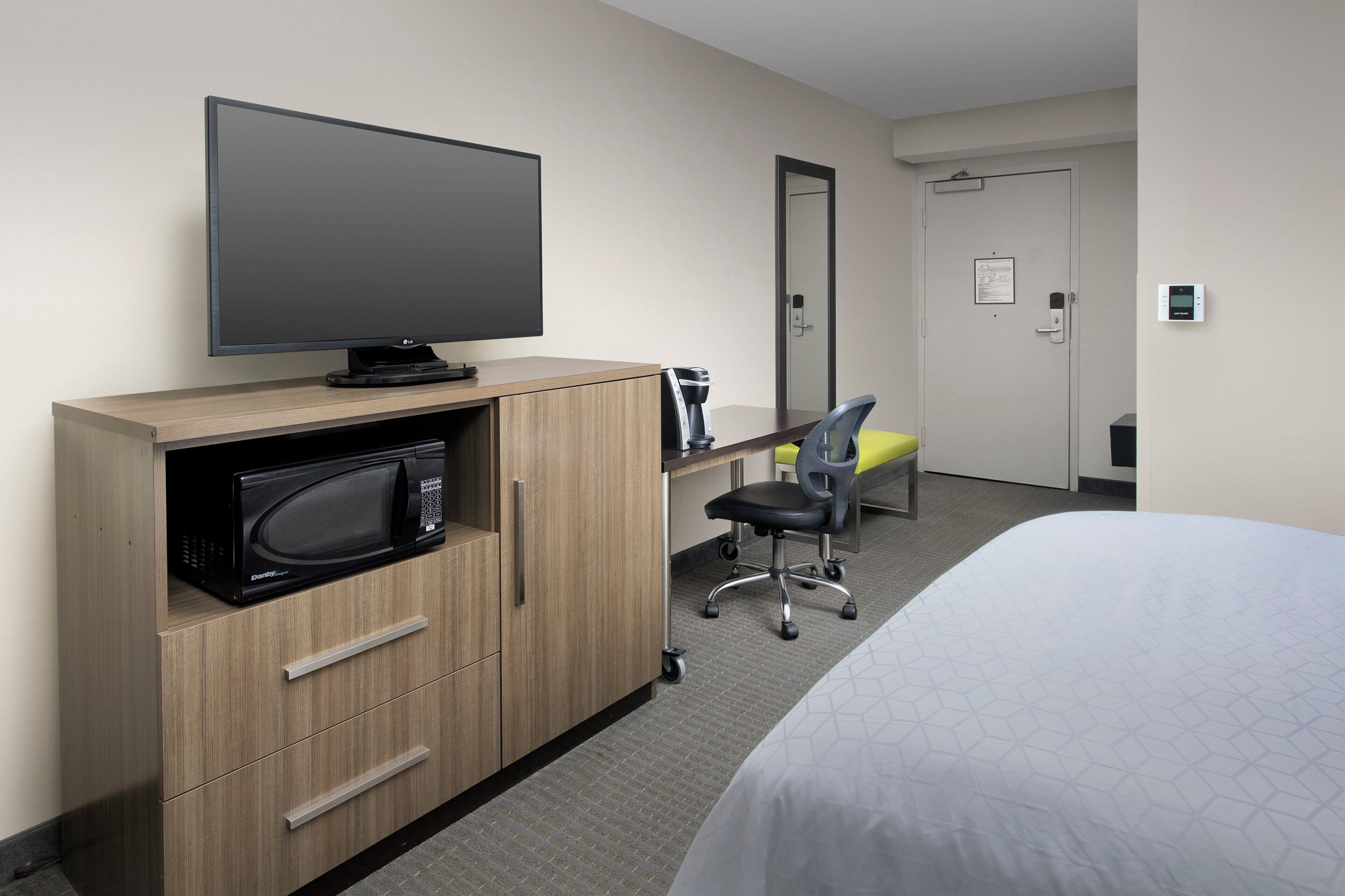 A hotel room with a bed, a TV on a wooden cabinet, a microwave, a desk with a chair, and a door with a lock.