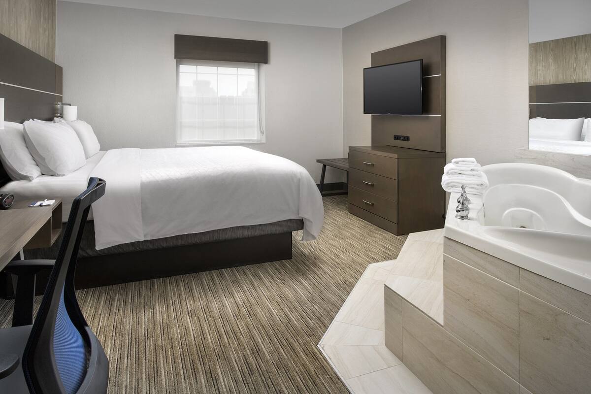 A modern hotel room with a large bed, a flat-screen TV, a bathroom with a bathtub, and a desk with a chair.