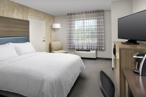 Habitación de hotel con una cama grande, un escritorio con televisor de pantalla plana, una silla, una lámpara de escritorio y una ventana con cortinas.