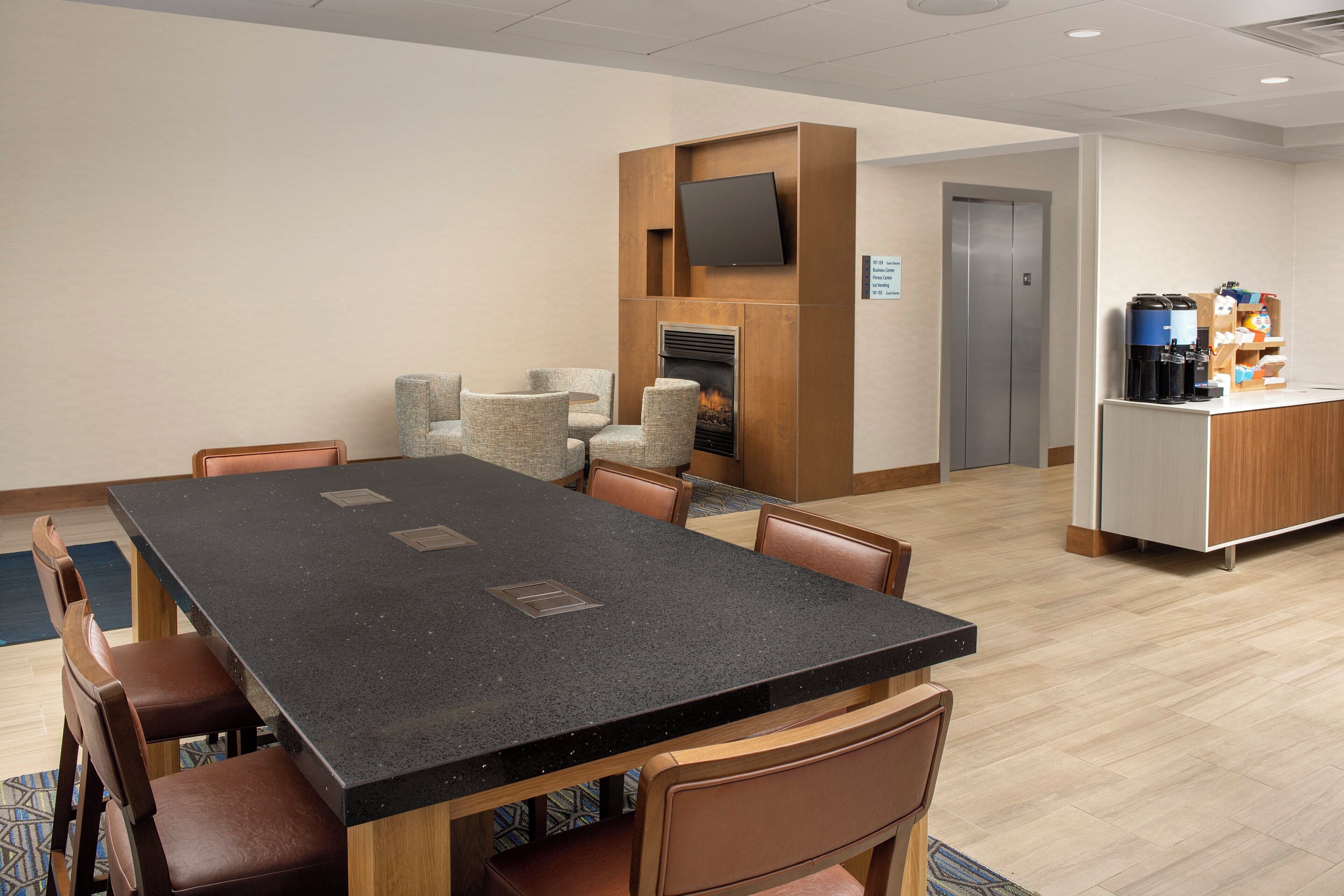 A hotel room with a large dining table, several chairs, a fireplace, a television, and a kitchenette area with coffee machines.