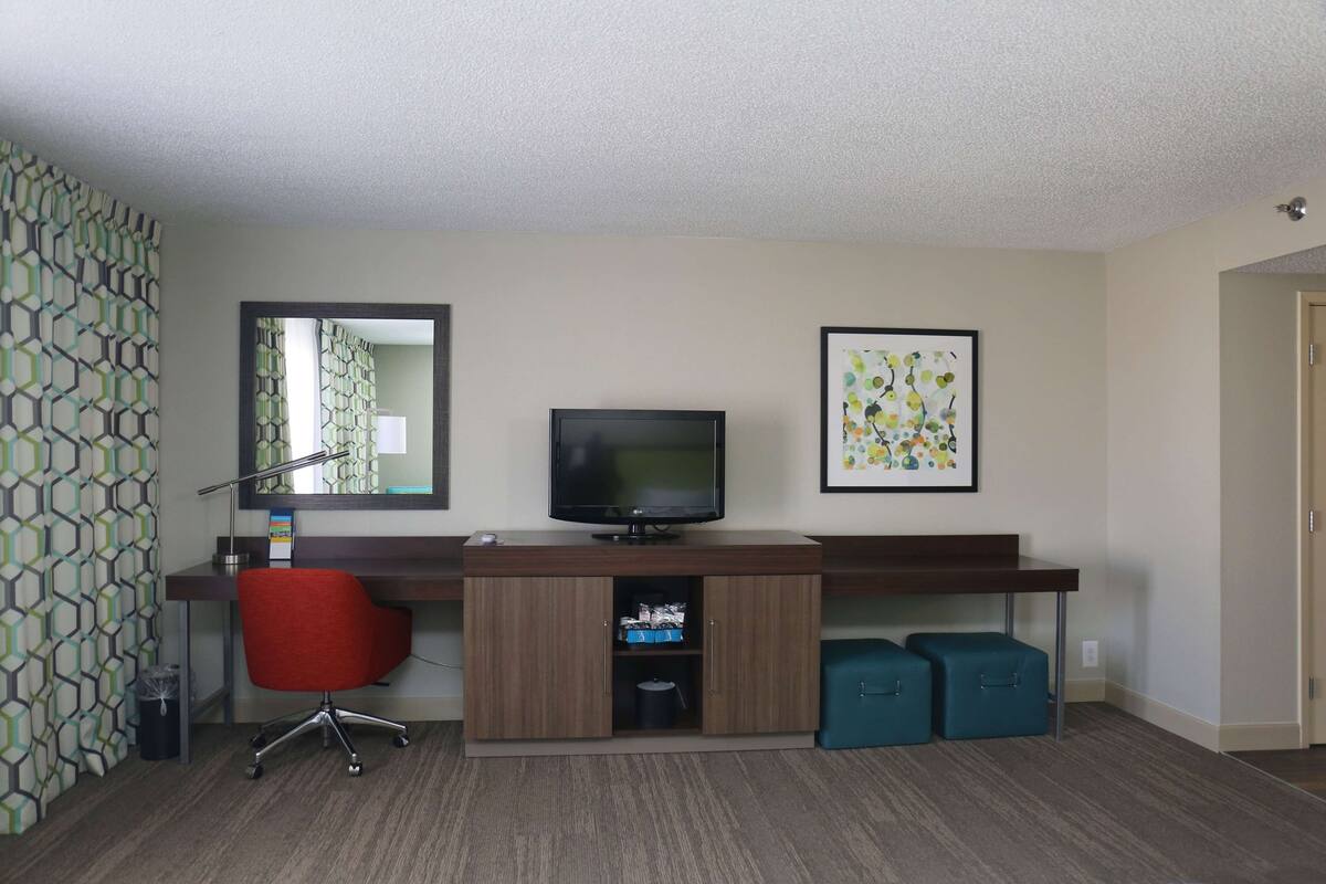 A hotel room with a wooden desk, a red office chair, a television, a framed artwork, and a small cabinet.