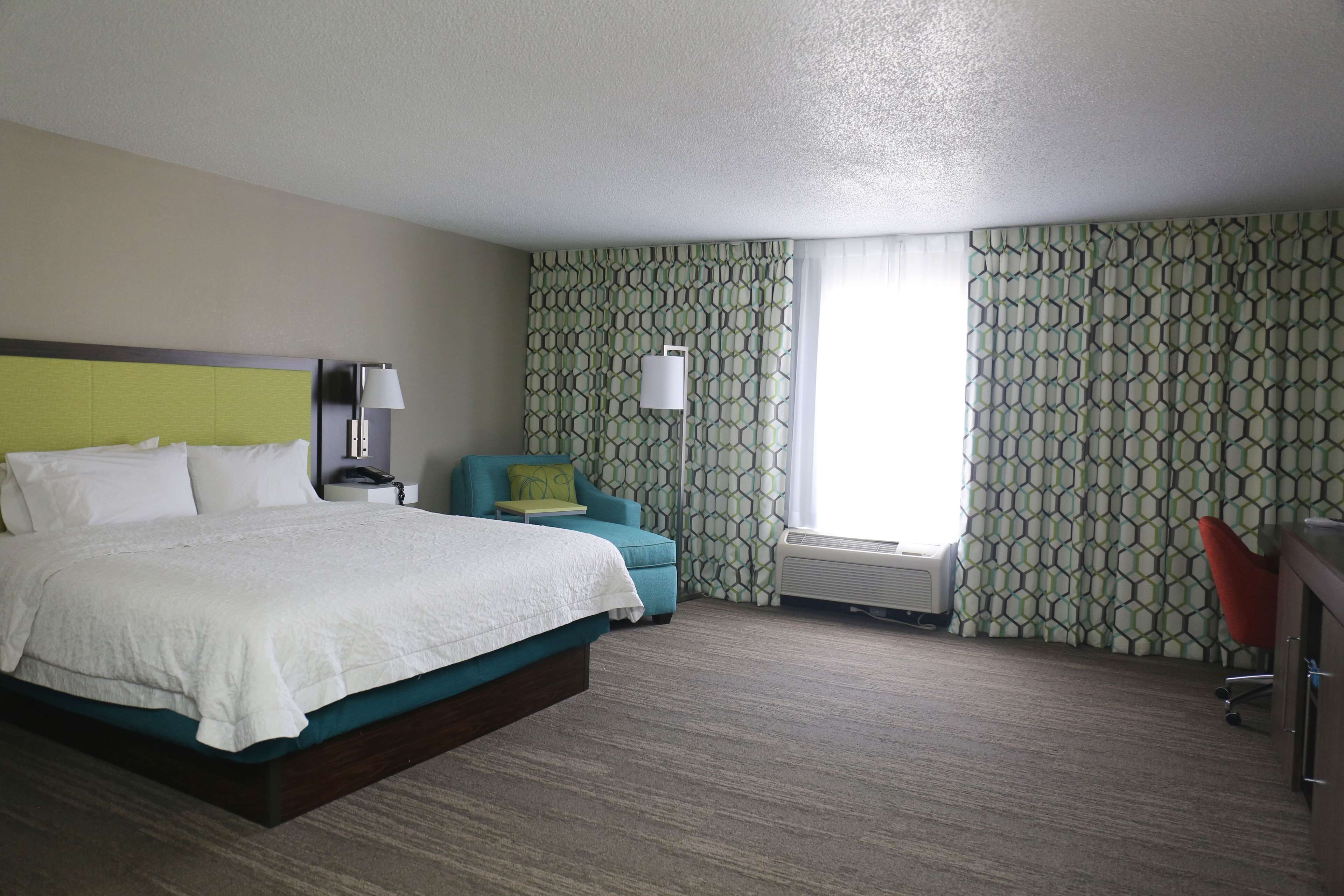 A hotel room with a large bed, a desk, a chair, and a window with patterned curtains.