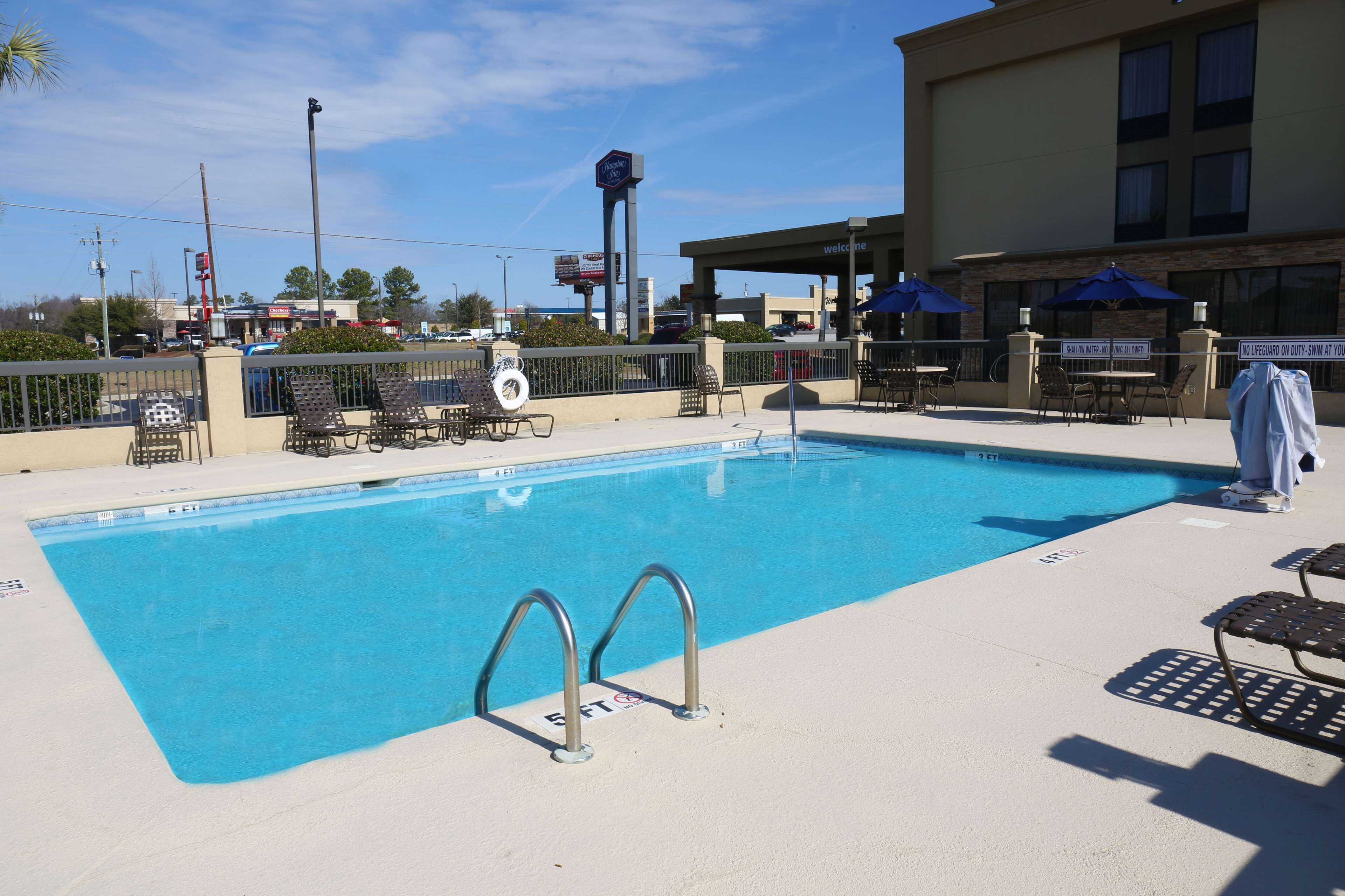 A swimming pool with lounge chairs and umbrellas, surrounded by a fence and a building with a 'Welcome' sign.