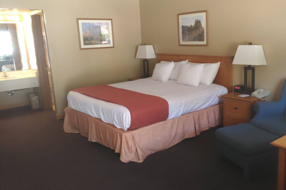 A hotel room with a bed, two bedside lamps, a chair, a table, and two framed pictures on the wall.