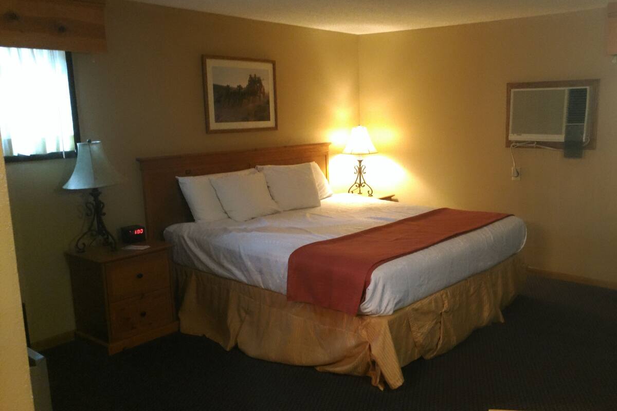 A hotel room with a bed, a bedside lamp, a nightstand, a clock, a picture on the wall, and an air conditioning unit.