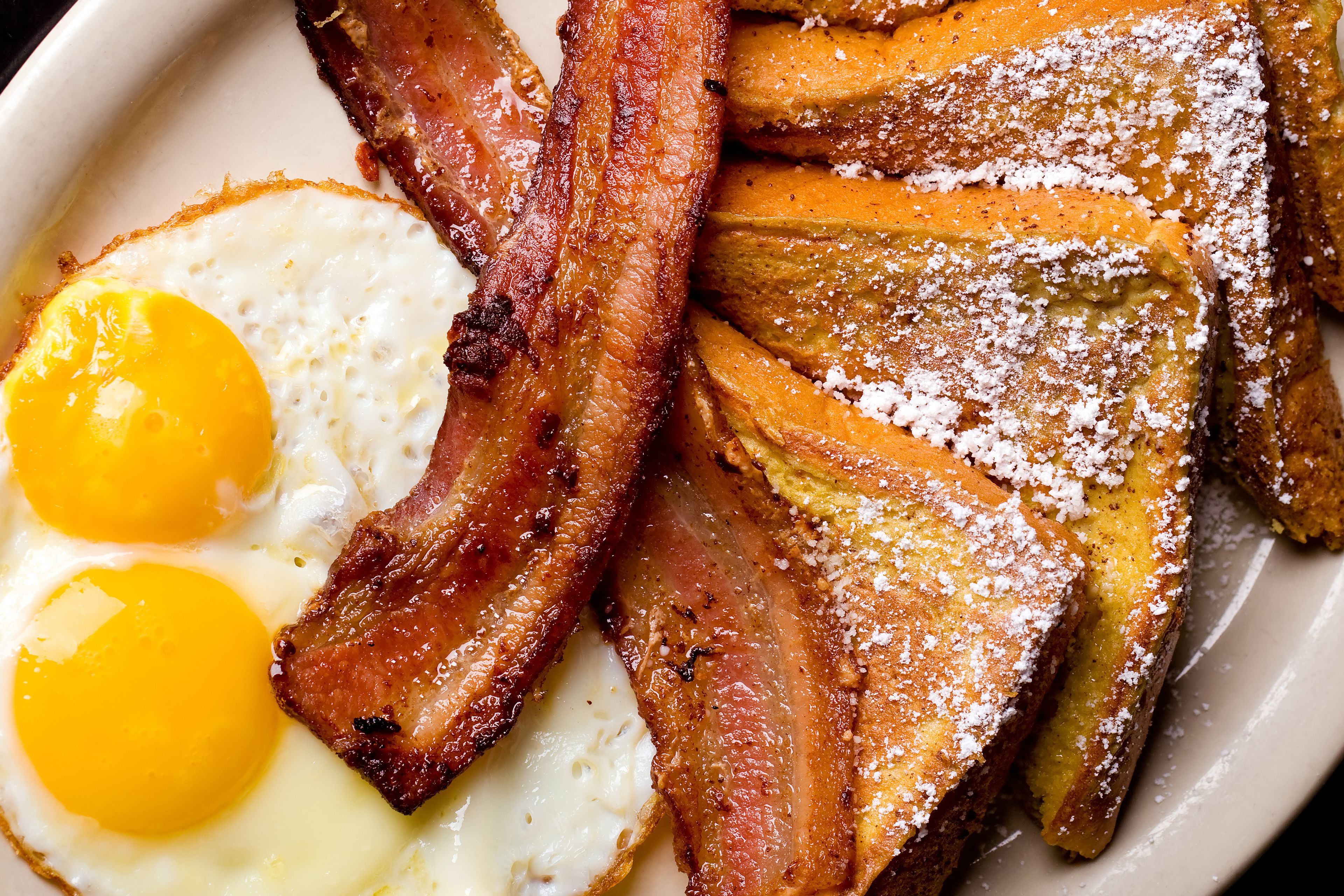 A plate with two fried eggs, crispy bacon, and French toast with powdered sugar.