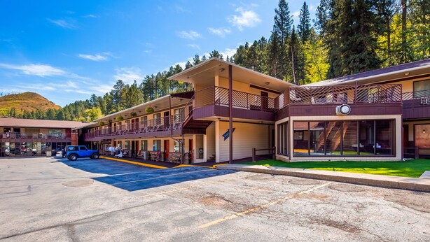 A two-story motel with a parking lot, outdoor seating, and a view of a forested hillside. - Deadwood Miners Hotel (Deadwood)