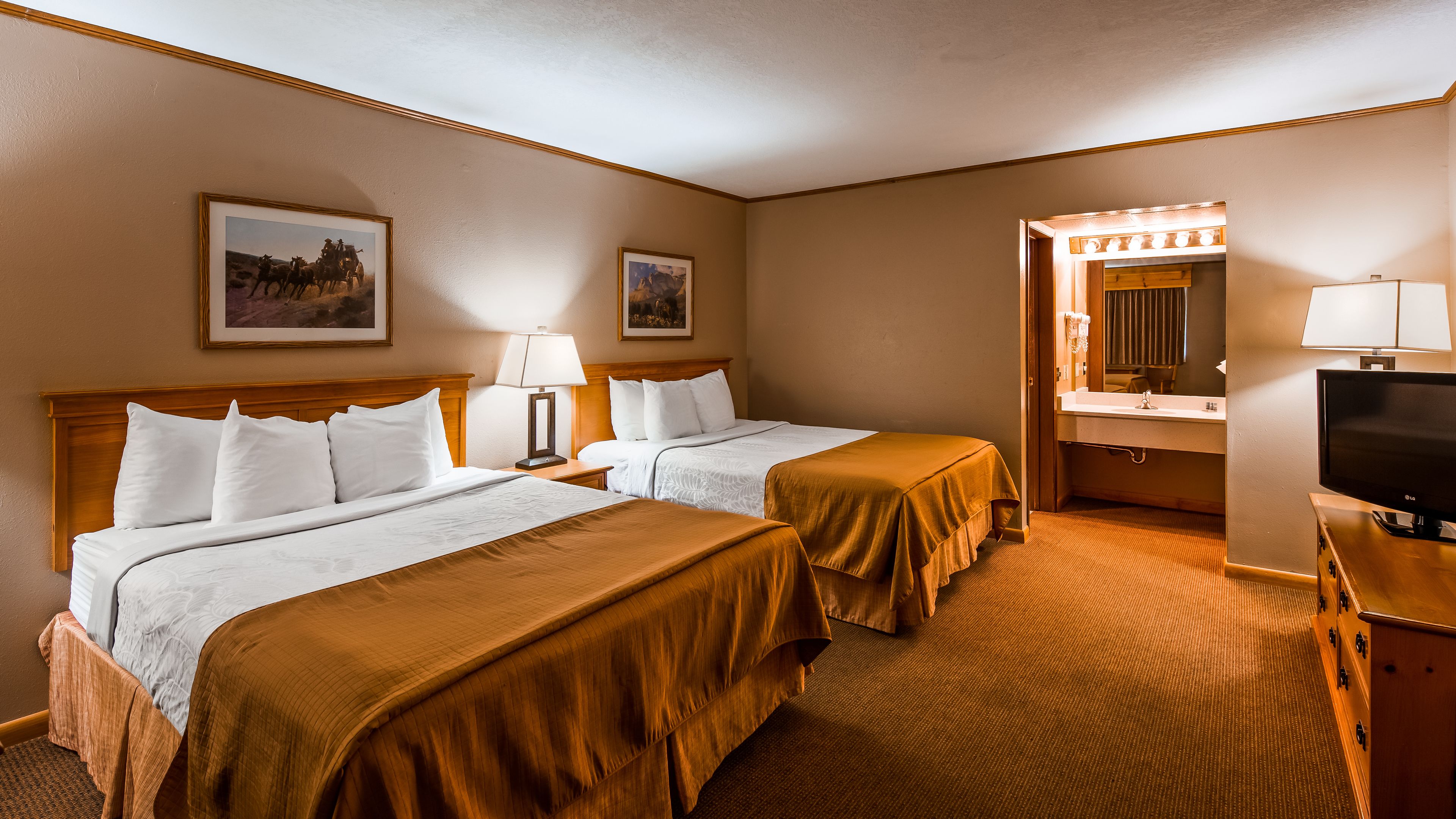A hotel room with two beds, a TV, a bathroom with a mirror and lights, and framed pictures on the wall.