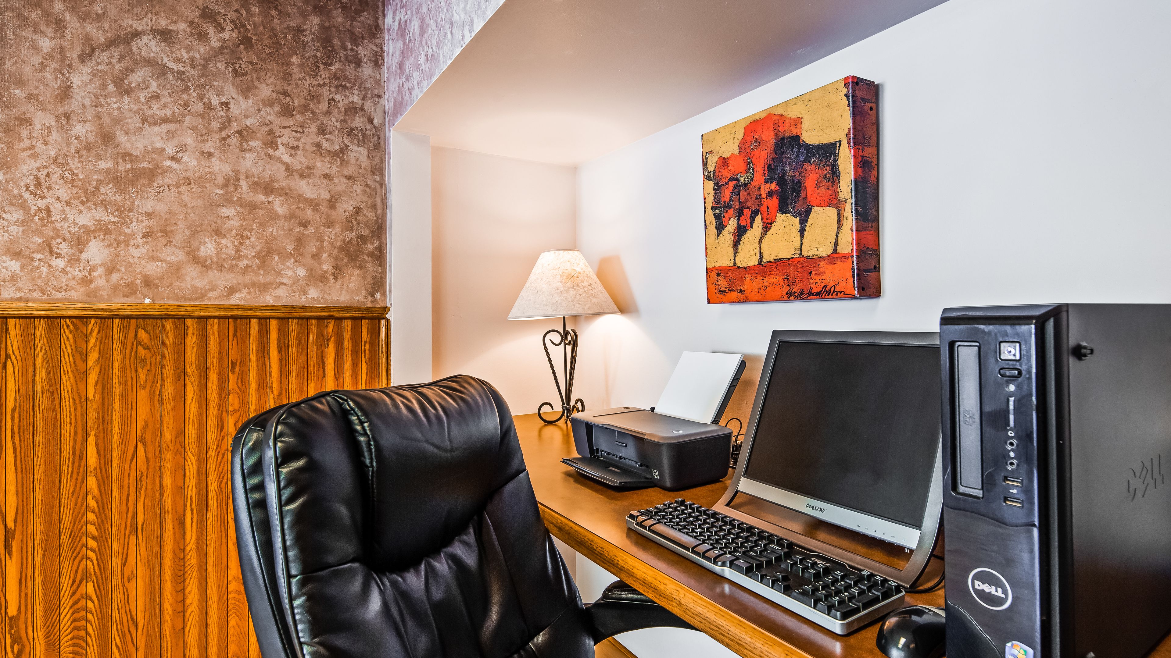 Une chambre d’hôtel avec un mur lambrissé en bois, un fauteuil en cuir noir, un bureau avec un ordinateur portable, un ordinateur de bureau, une lampe et un tableau accroché au mur.