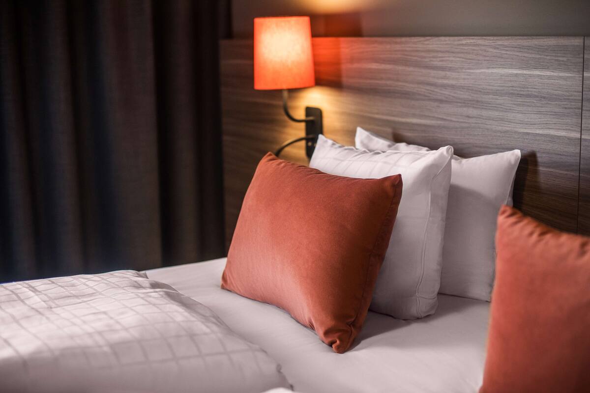 A neatly made bed with white and brown pillows, a wooden headboard, and a wall-mounted lamp.