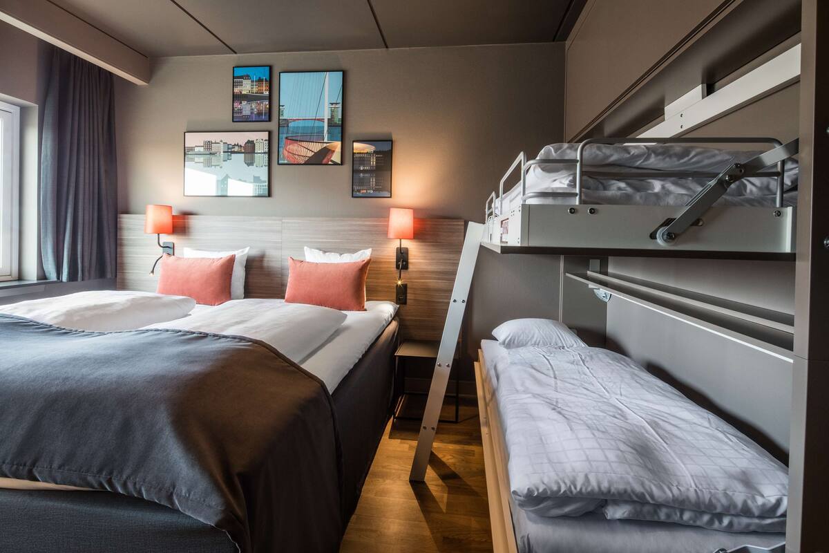 A hotel room with two beds, a bunk bed, a wooden headboard, and wall-mounted lamps.
