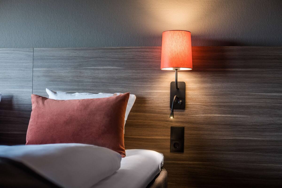 A hotel room with a bed, a red pillow, a wall-mounted lamp, and a control panel.