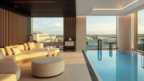 A modern indoor swimming pool with a sofa and ottoman, large windows, and a city view.