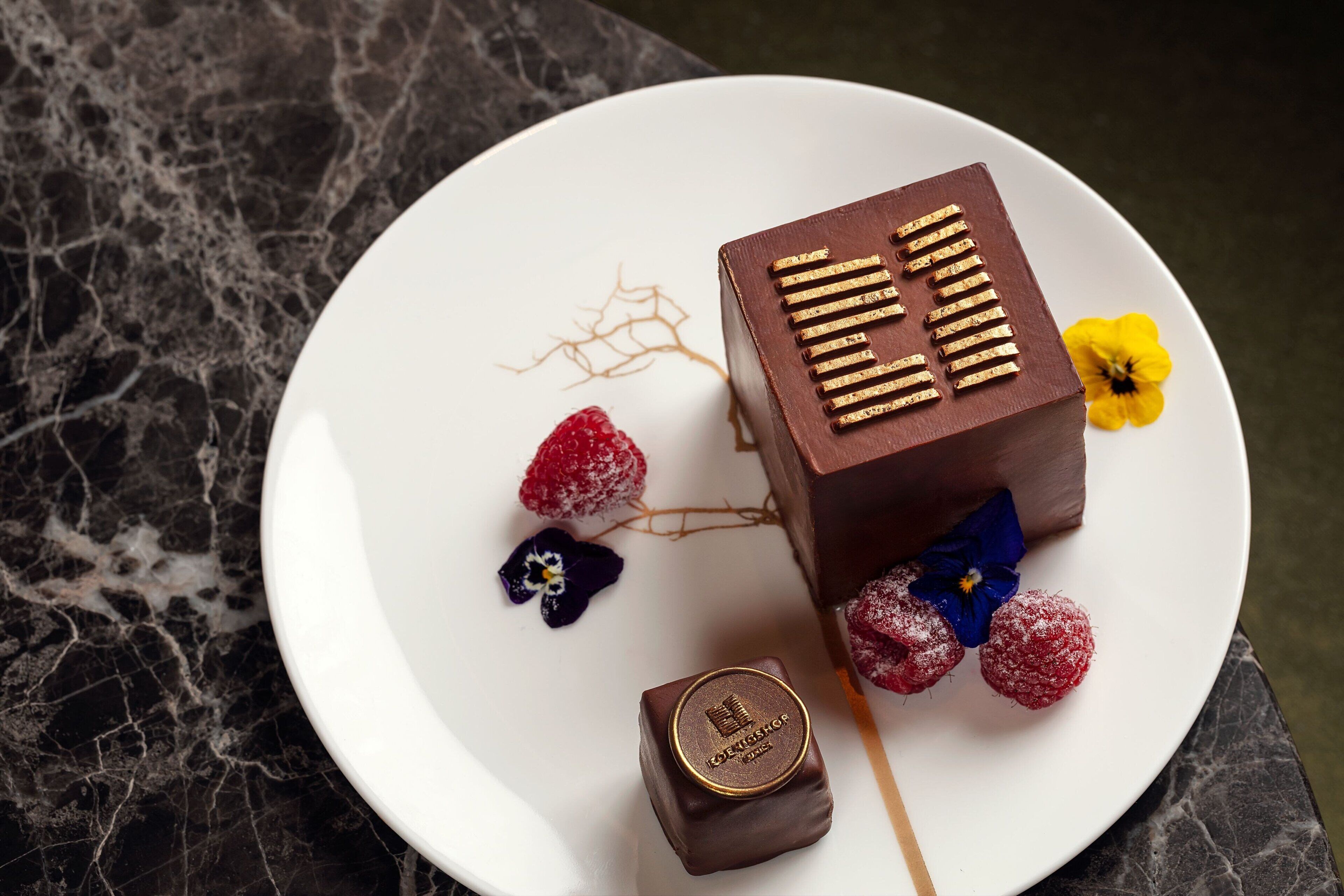 Un postre sobre un plato blanco con chocolate, frambuesas y flores.